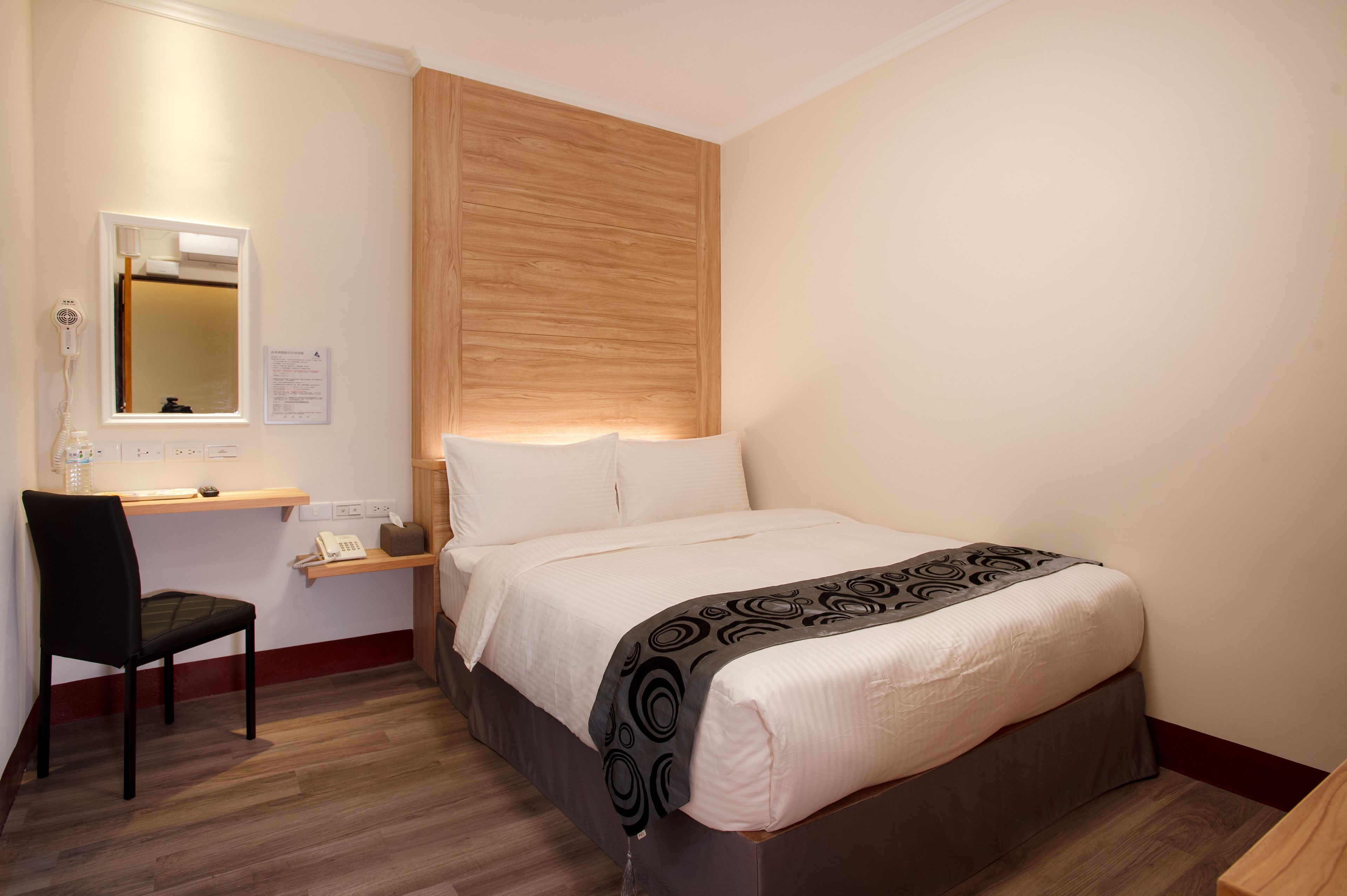 Una habitación de hotel con cama, escritorio, silla, espejo y cabecera de madera.