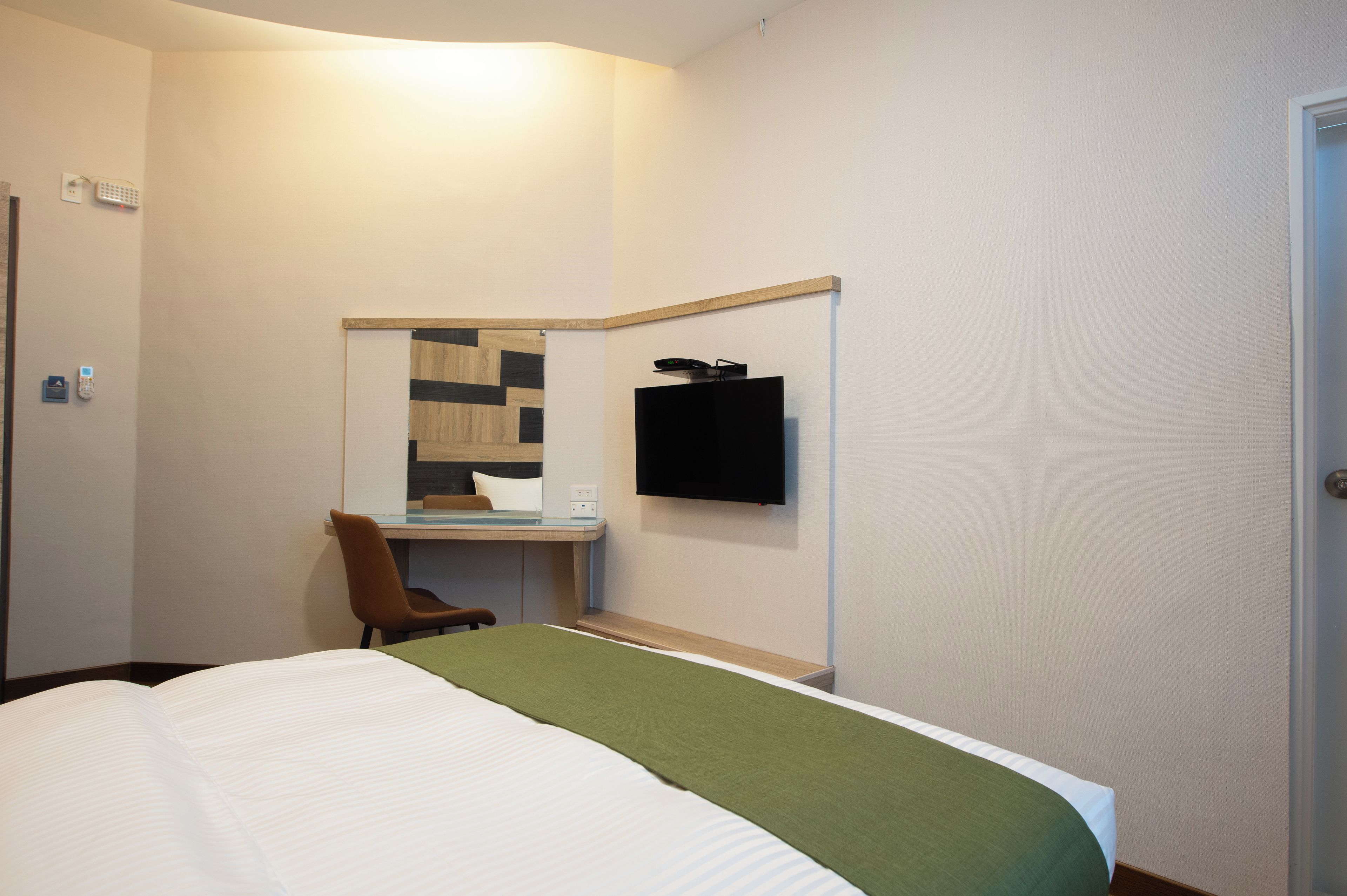 A hotel room with a bed, a desk, a chair, a flat-screen TV mounted on the wall, and a door.