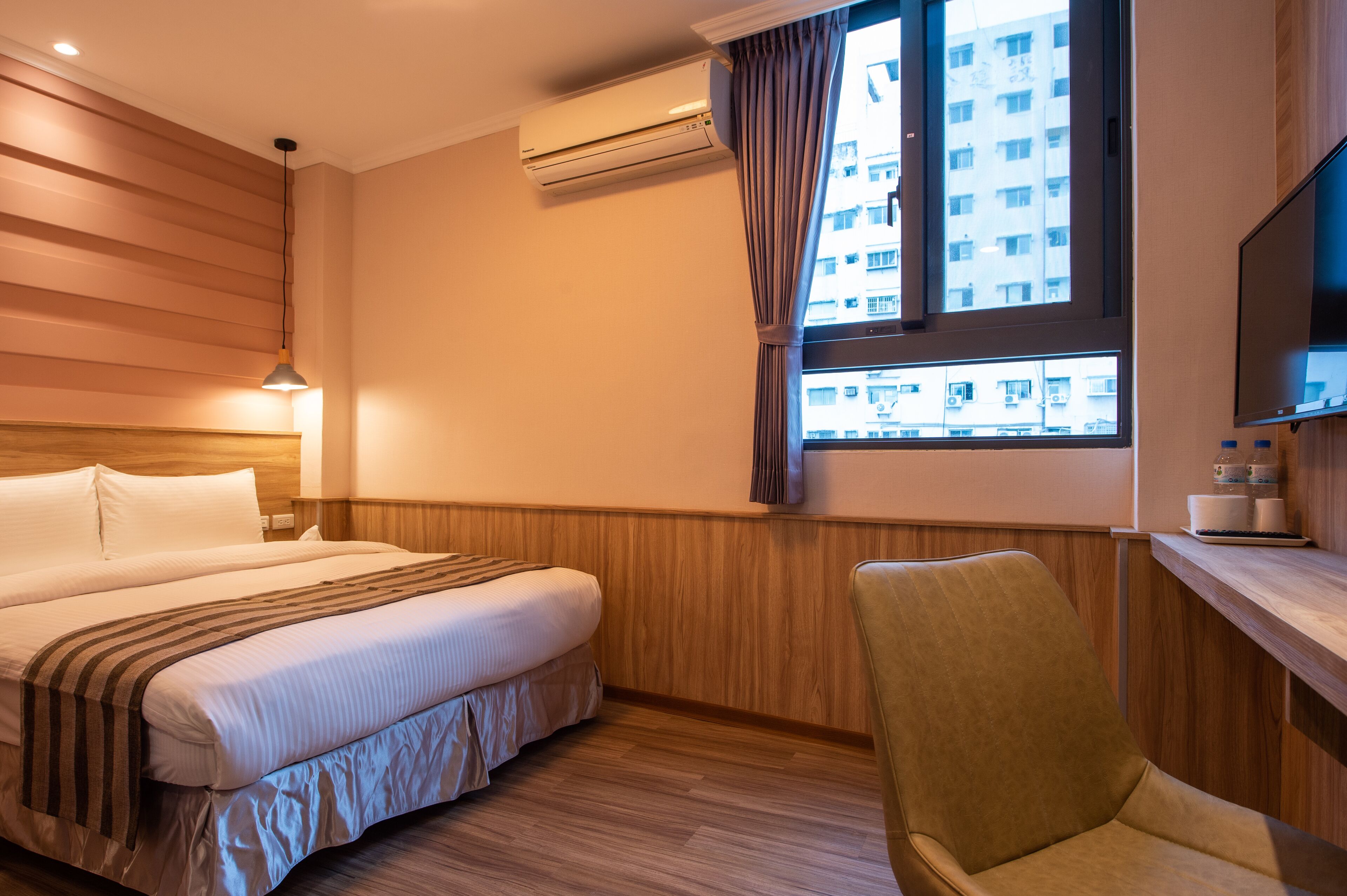 A hotel room with a bed, a chair, a TV, and a window with a view of buildings.