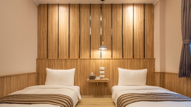 Habitación de hotel con dos camas, paneles de madera en las paredes, una mesita de noche con lámpara y un pequeño estante con objetos.