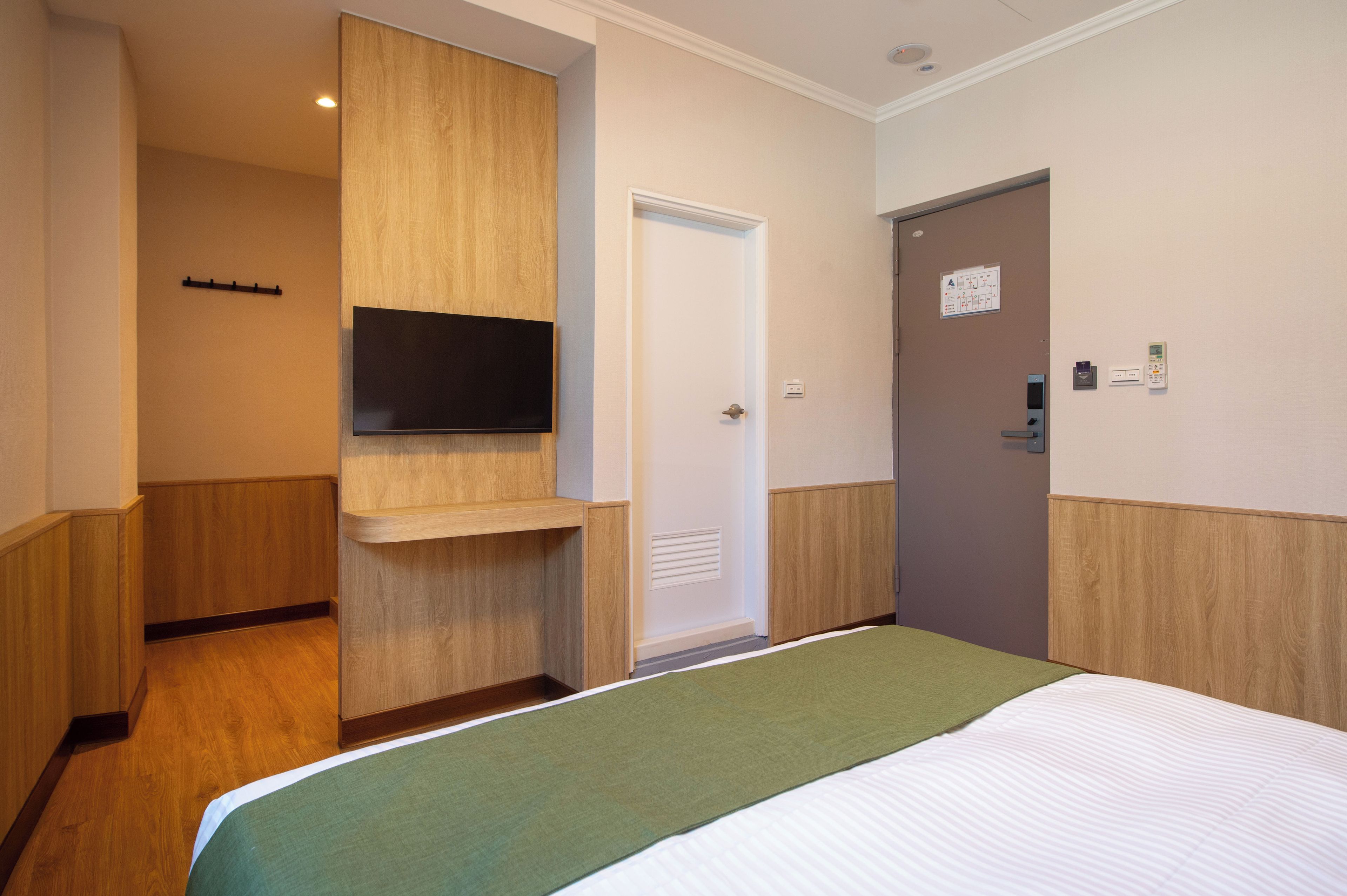 A hotel room with a bed, a mounted TV, a door with a lock, and a wall-mounted thermostat.