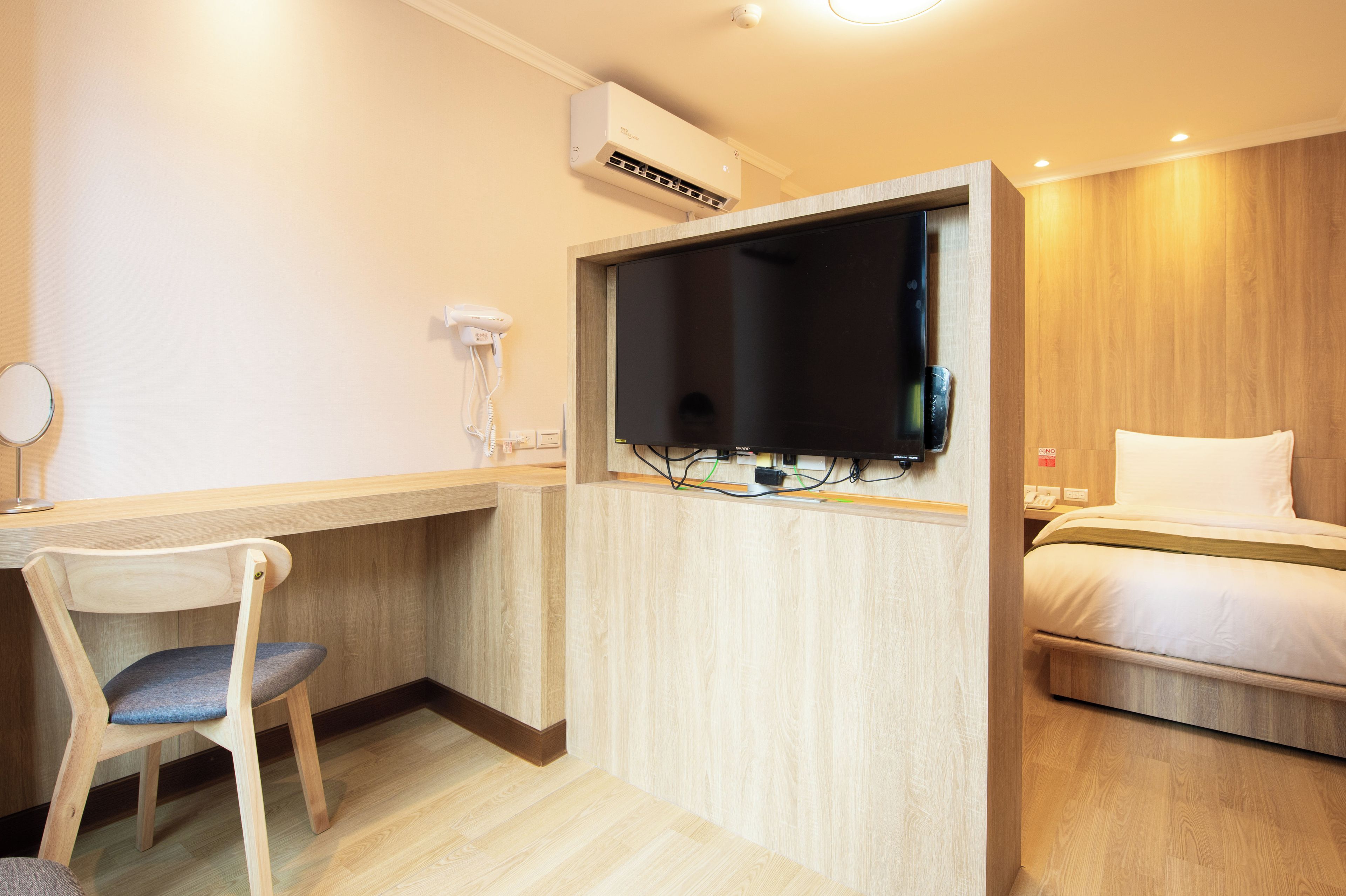 A hotel room with a bed, a desk with a chair, a television mounted on a wooden partition, and an air conditioning unit on the wall.