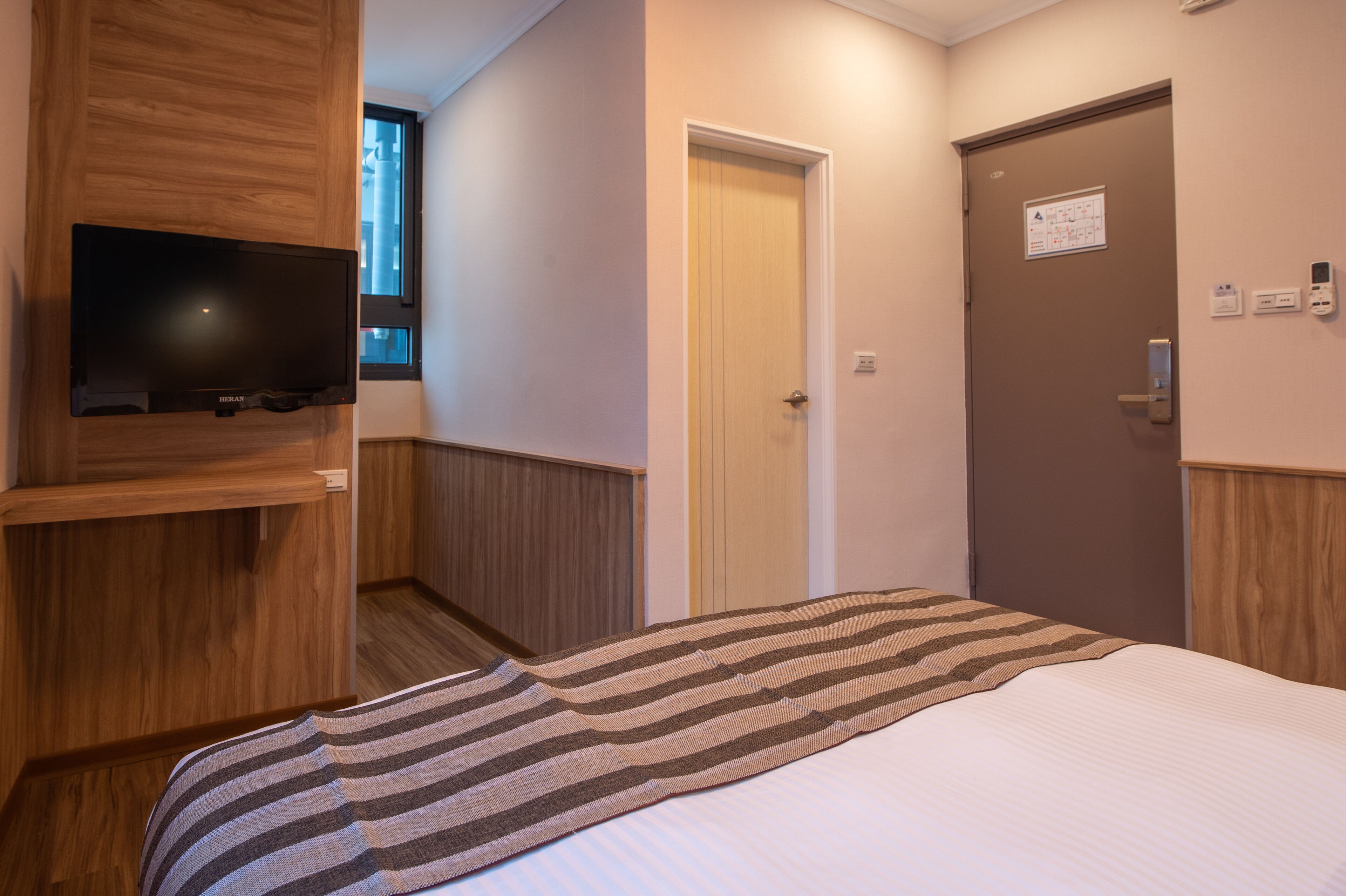 Una camera d'albergo con un letto, una televisione fissata a un pannello di legno, una porta con un cartello e una finestra.