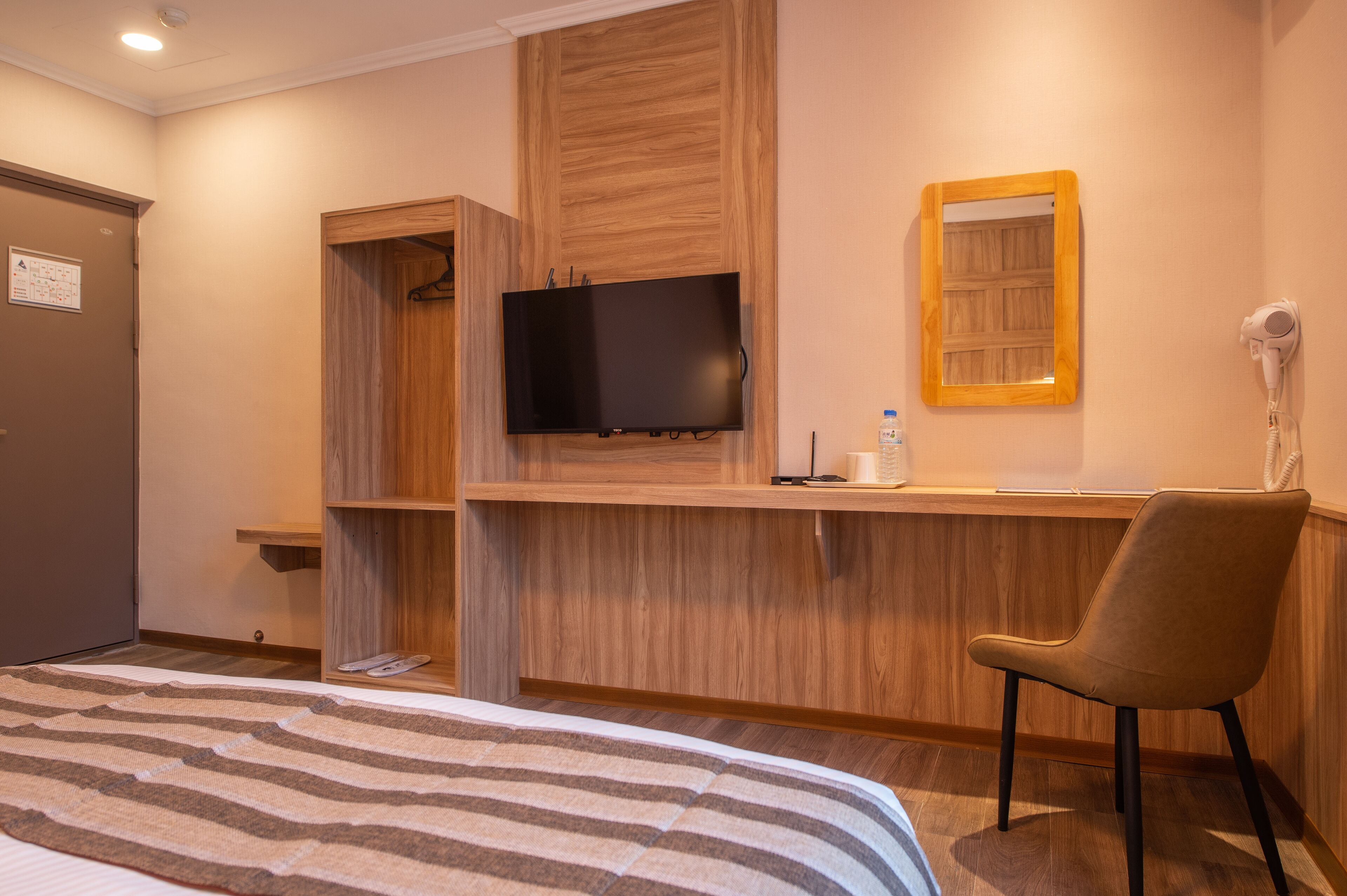 A hotel room with a bed, a TV mounted on a wooden panel, a chair, a mirror, and a hairdryer on the wall.