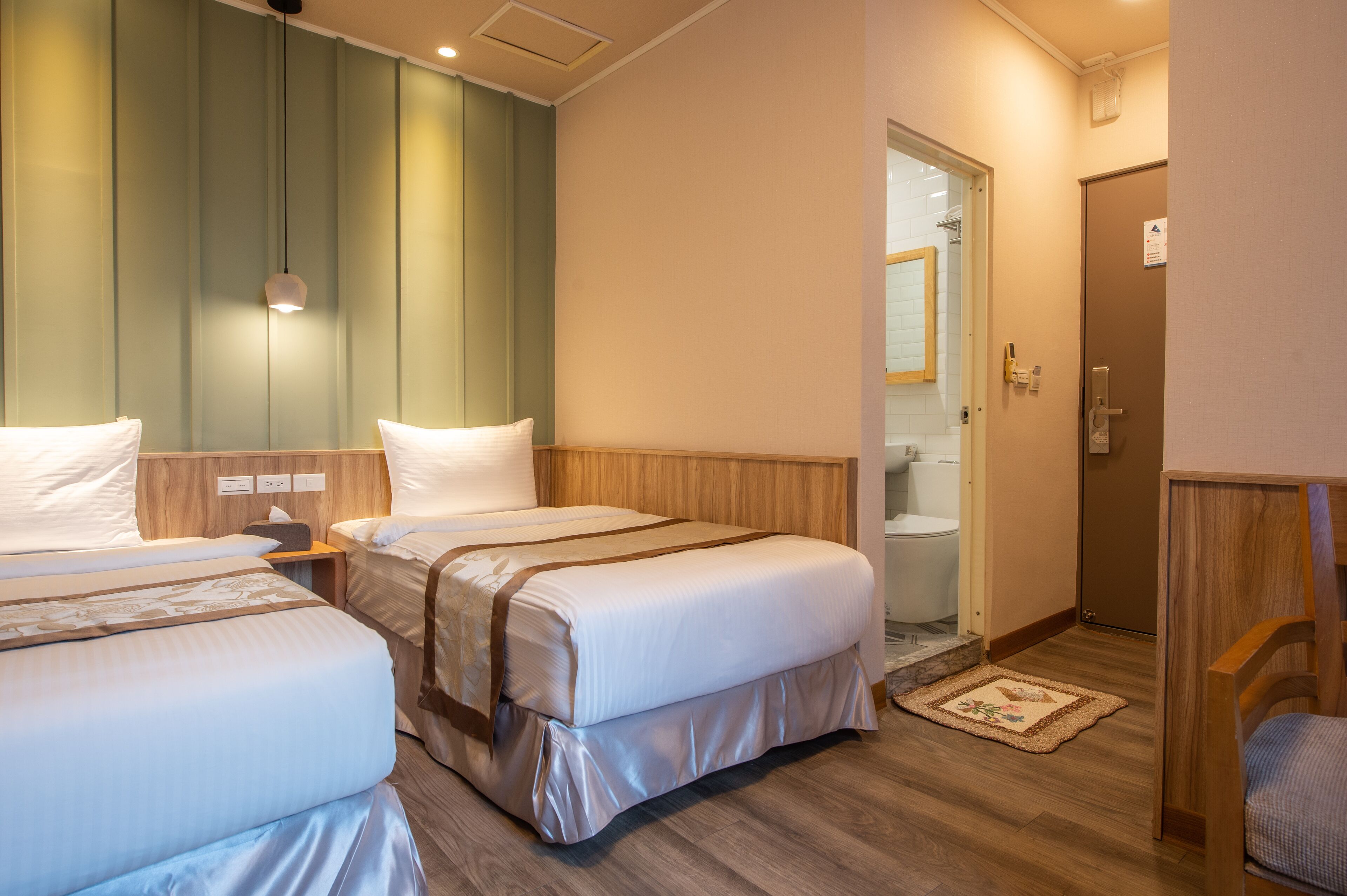 Habitación de hotel con dos camas, cabecero de madera, mesita de noche y puerta al baño.