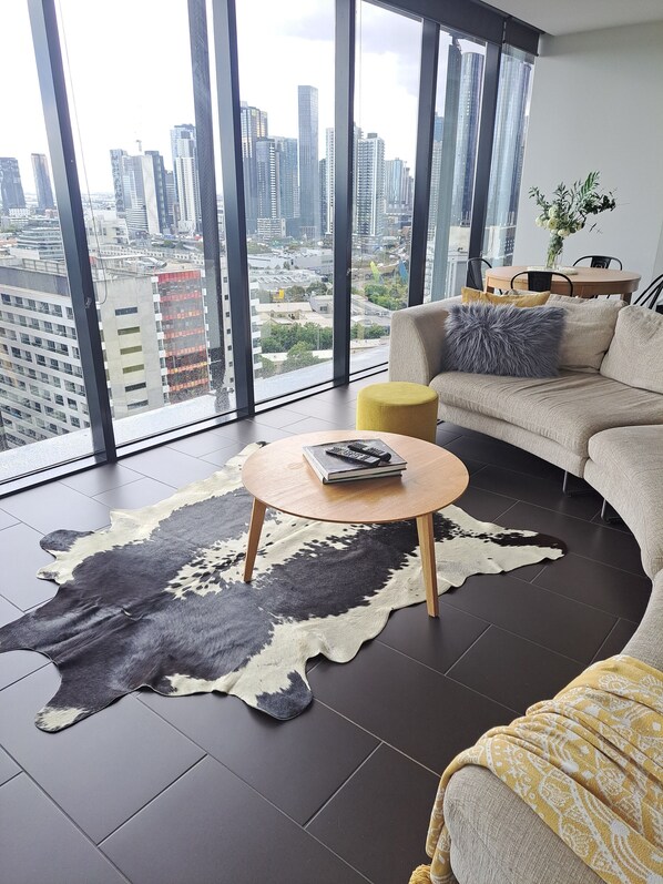 A modern living room with a large city view, a leather rug, a round coffee table, and a sofa with cushions. - Xanadu Apartments (Southbank)