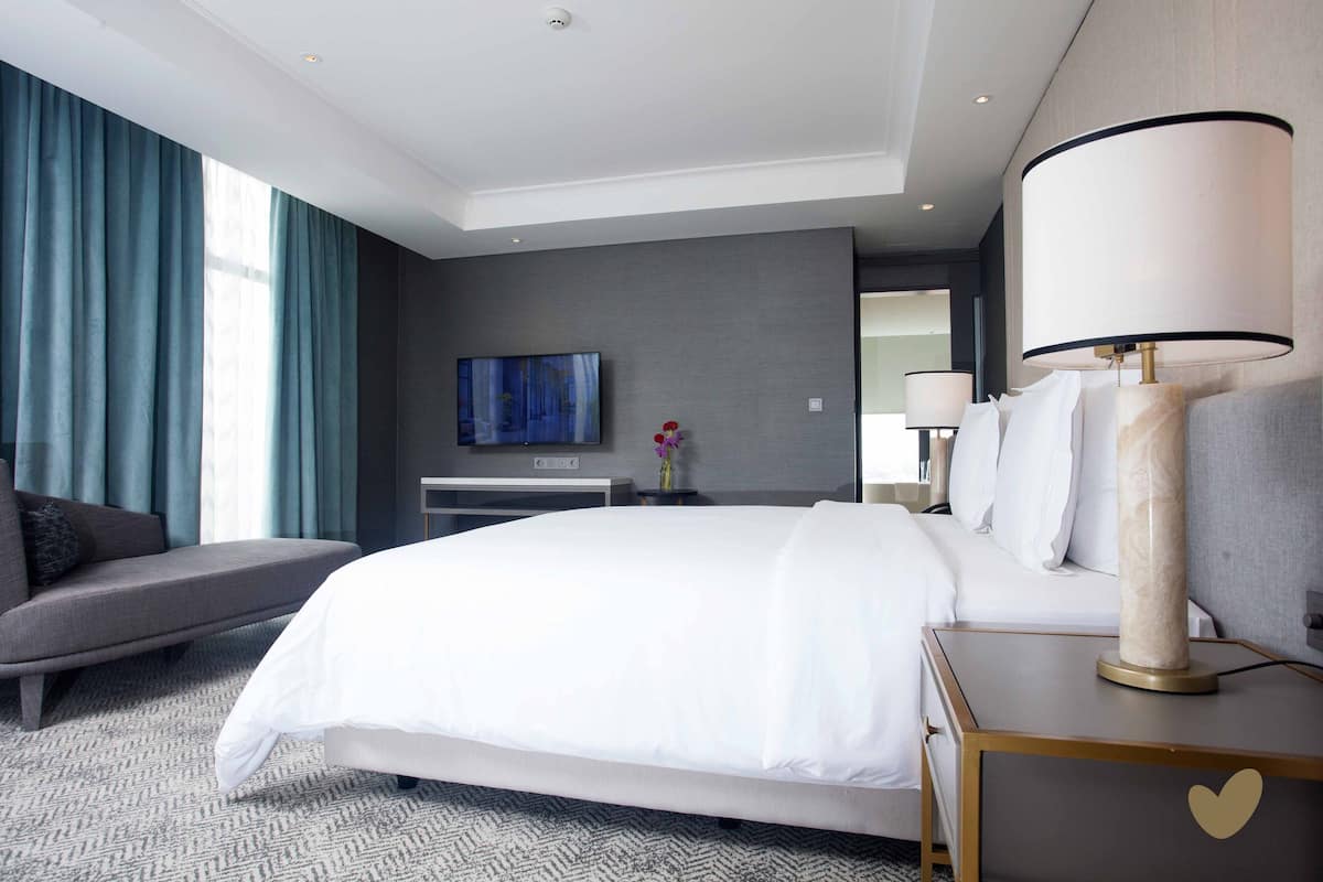 A modern hotel room with a large bed, a sofa, a TV, and a bedside table with a lamp.