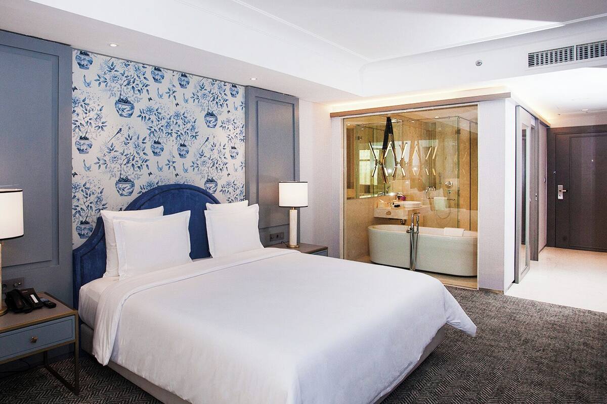 A hotel room with a large bed, two bedside lamps, a nightstand, a bathroom with a glass shower enclosure, and a blue floral wallpaper.
