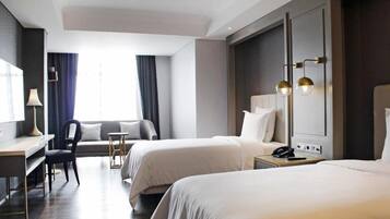 Quarto de hotel com duas camas, uma escrivaninha, uma cadeira, um abajur e uma janela com cortinas.