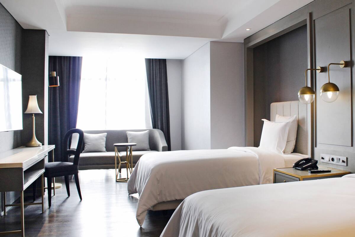 A hotel room with two beds, a desk, a chair, a small table, and a nightstand with a phone.