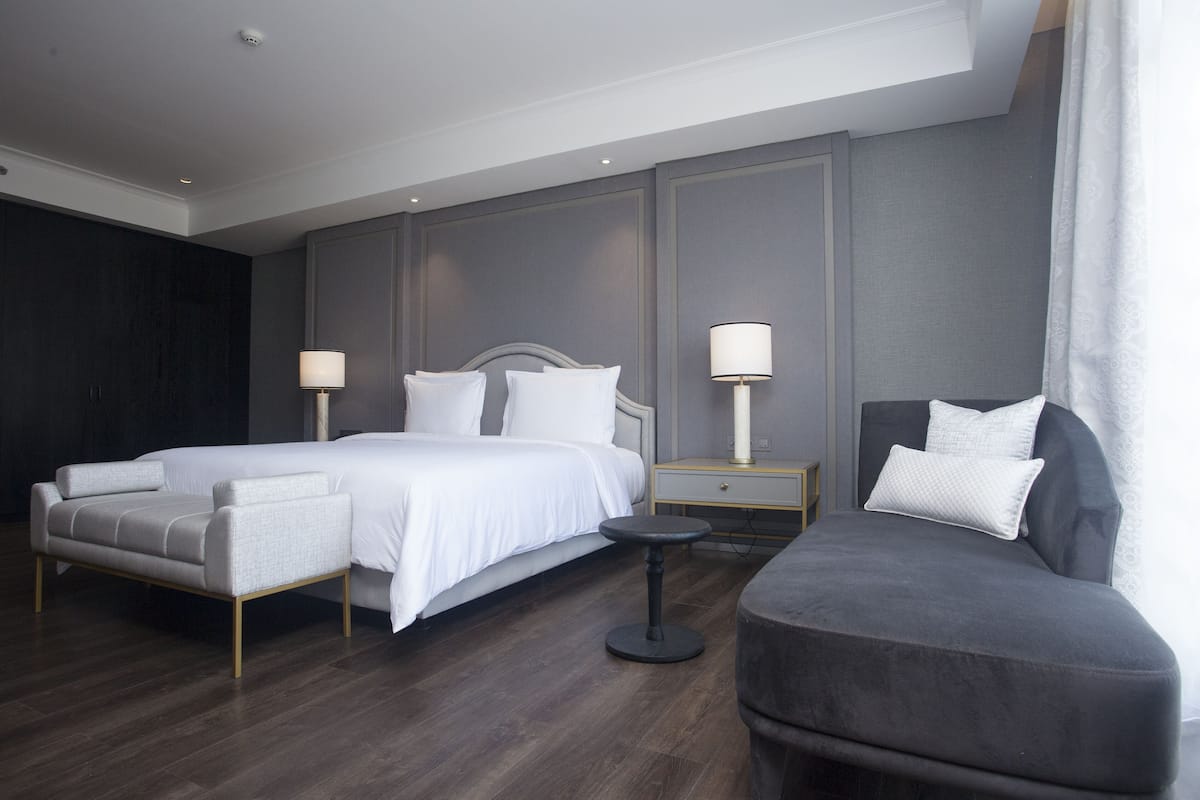 A modern hotel room with a large bed, two bedside tables with lamps, a grey armchair, and a small round table.