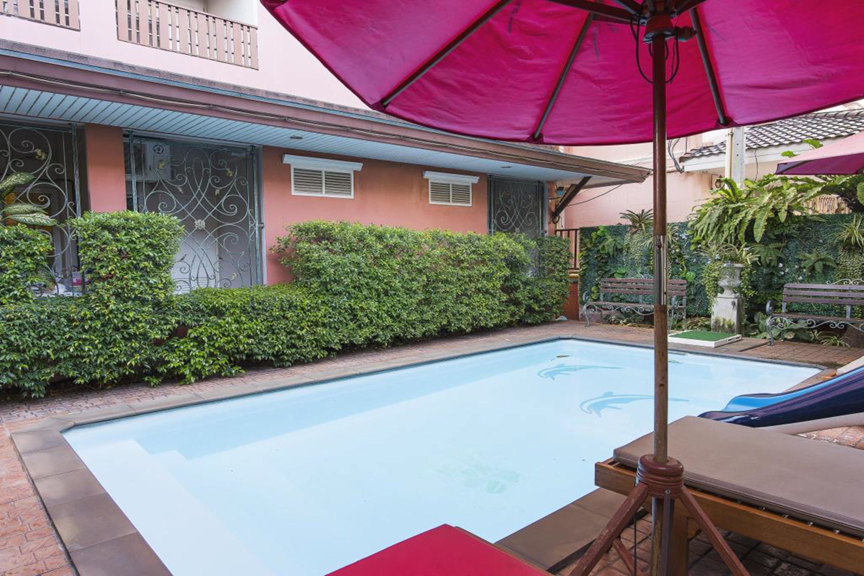 A swimming pool with lounge chairs and a large umbrella, surrounded by a well-maintained garden and a building with decorative iron gates.