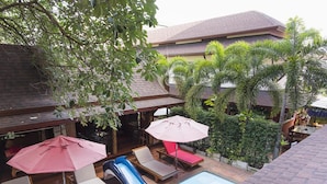 An outdoor swimming pool area with lounge chairs and umbrellas, surrounded by lush greenery and a building with a tiled roof. - Orchid Resort (Bangkok)