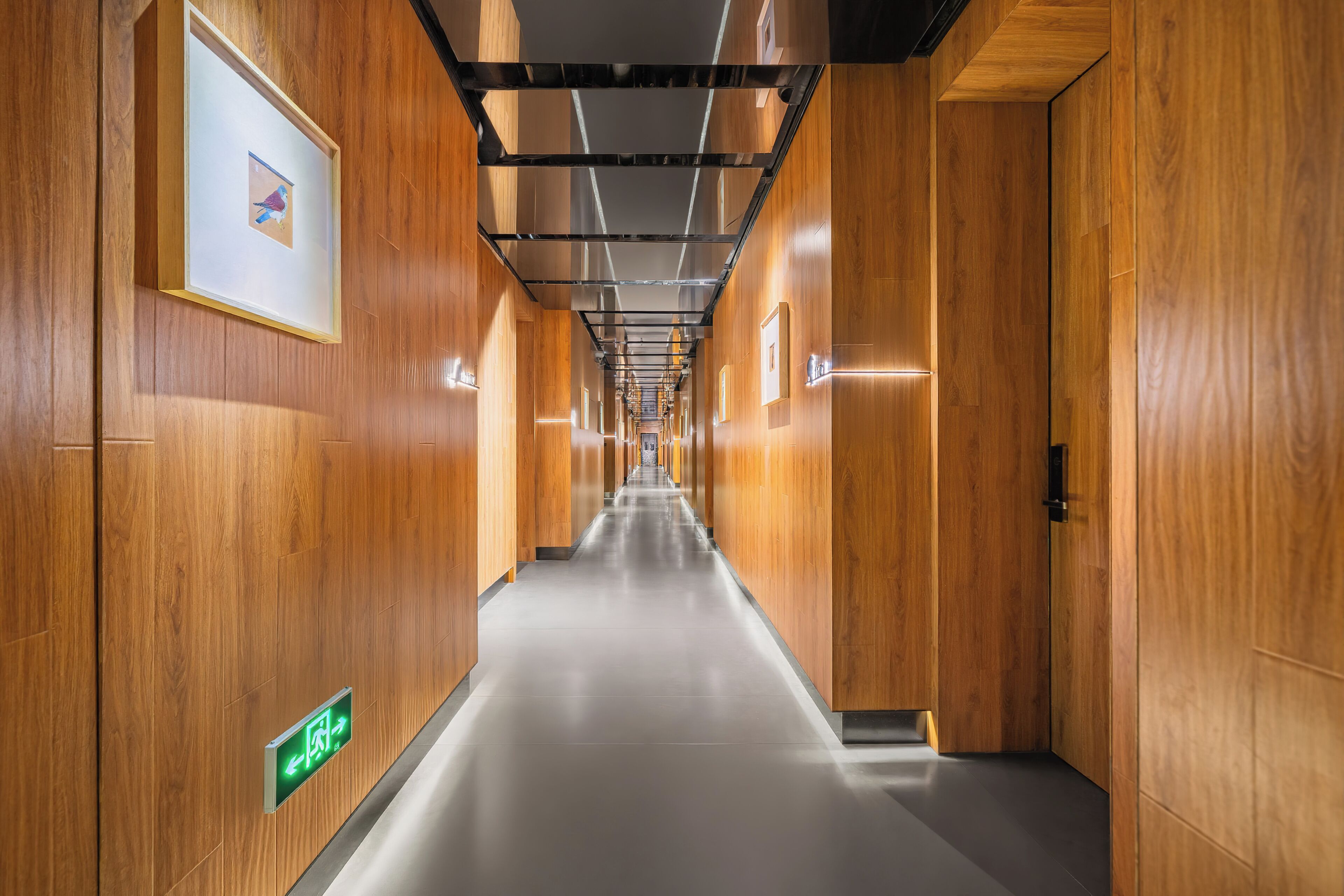 A long hallway with wooden walls and a tiled floor, featuring a green exit sign and framed artwork on the left.