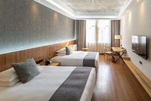 A hotel room with two beds, a desk, a chair, a TV, and a window with curtains.