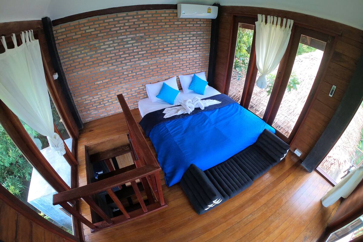 A room with a brick wall, a bed with blue bedding, a wooden chair, and a window.
