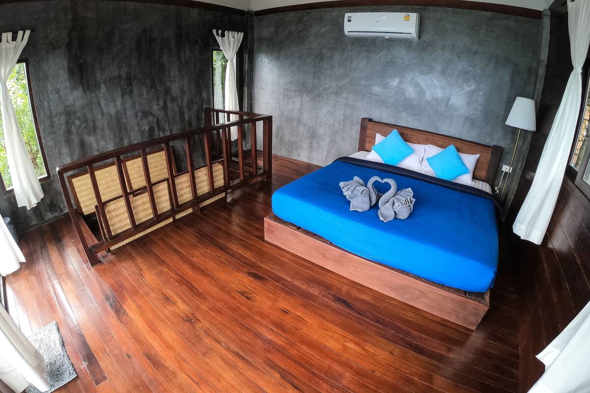A bedroom with a wooden floor, a bed with blue bedding, and a window with curtains.