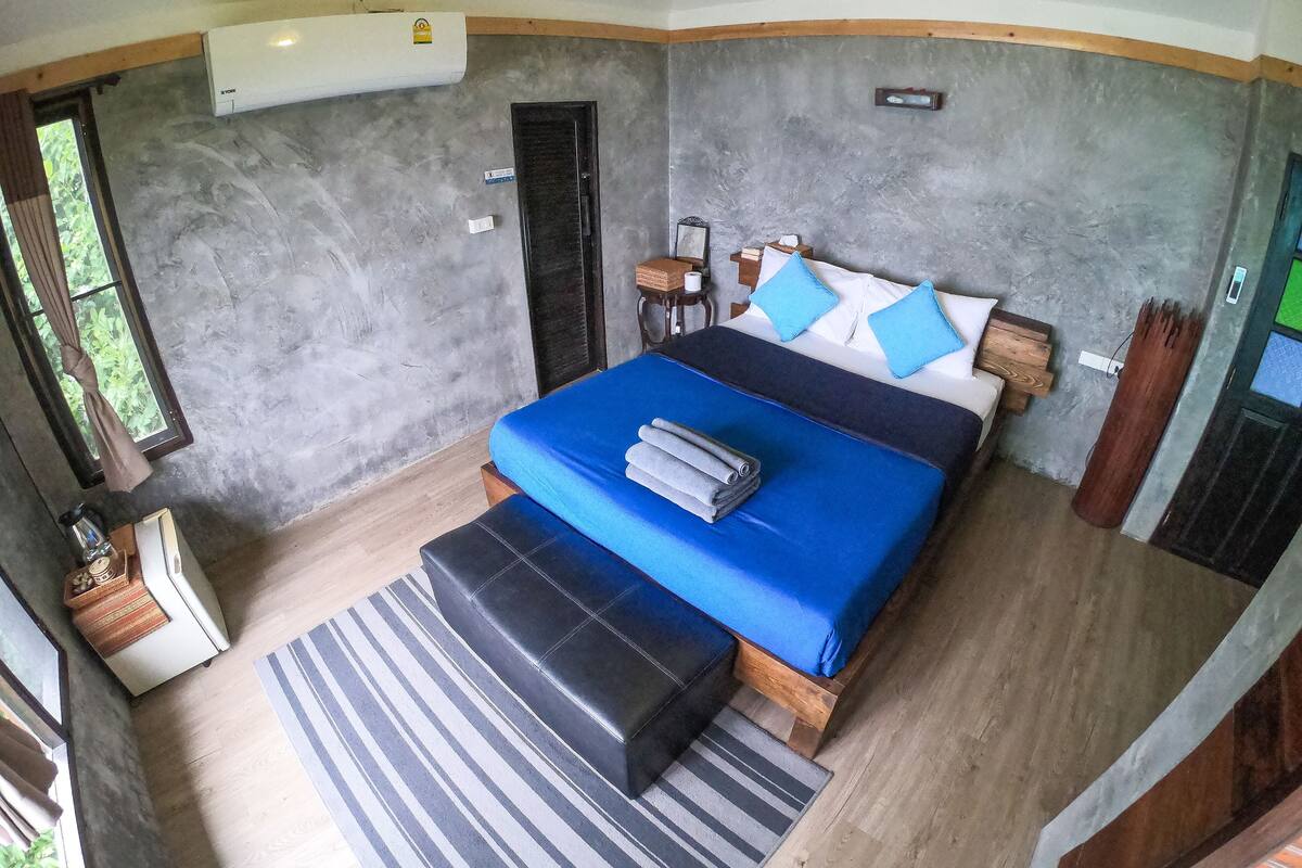 A bedroom with a large bed, a small bedside table, a nightstand, a window with curtains, and a wall-mounted air conditioner.