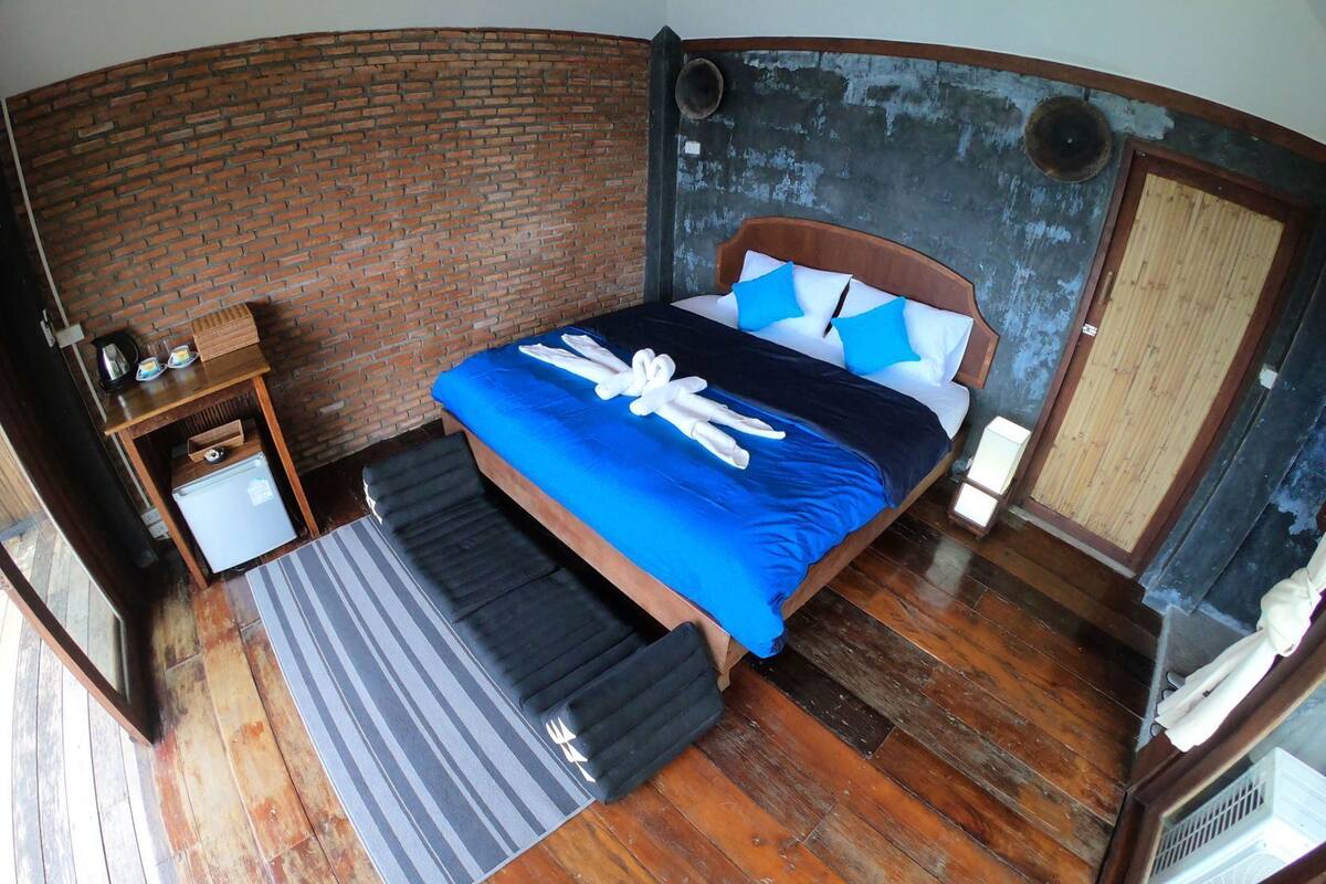 A room with a brick wall, a bed with blue bedding, a small table with a kettle, and a wooden bench.