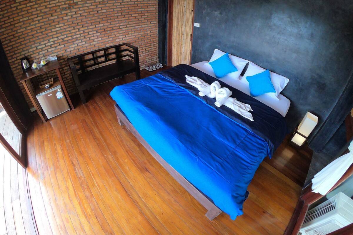 A room with a brick wall, a bed with blue bedding, a wooden bench, a small table with a refrigerator, and a door.