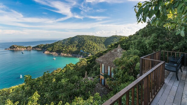 A wooden deck with a railing overlooking a turquoise sea and a lush green hillside.