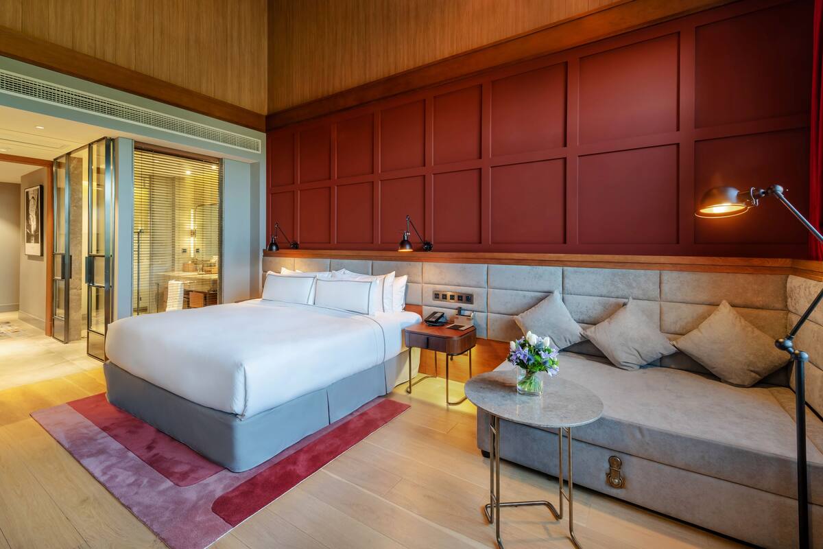A hotel room with a large bed, a sofa, a small table with a vase, and a nightstand with a lamp.
