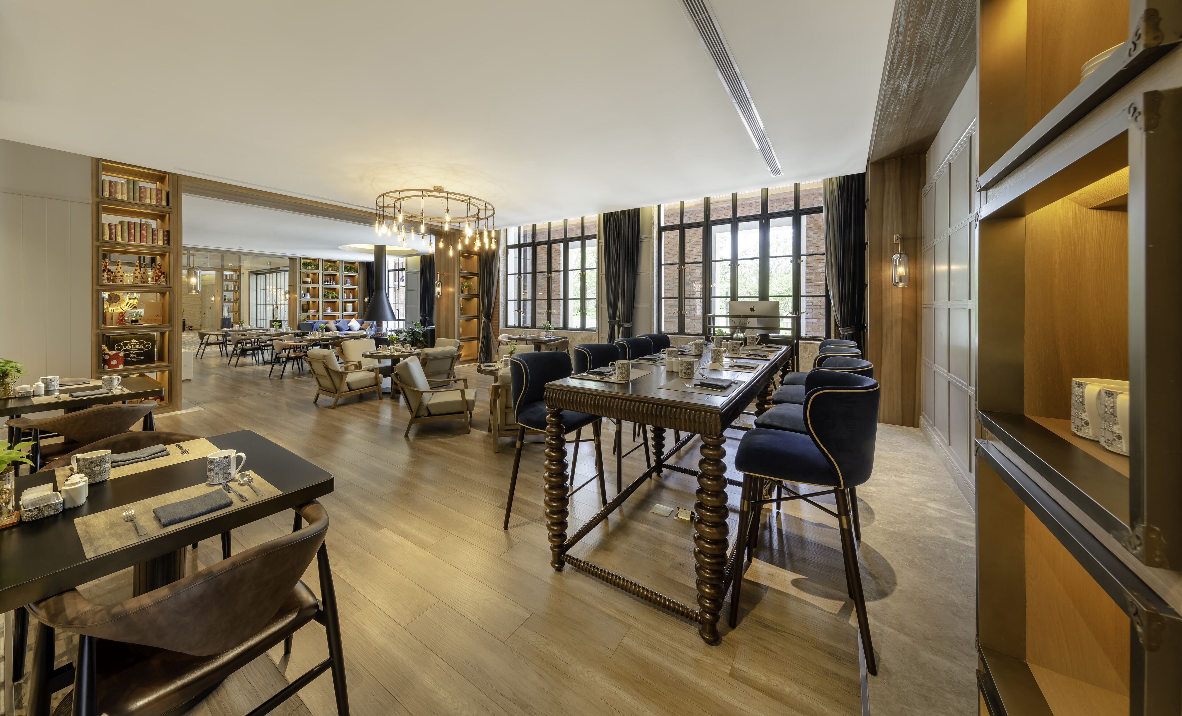 A spacious hotel lobby with a central seating area, a bookshelf, and a large window providing natural light.