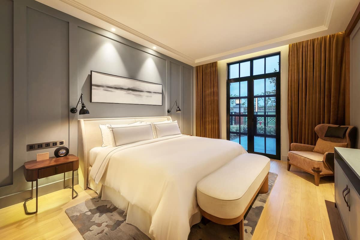 A modern hotel room with a large bed, a bedside table with a radio, a chair, and a view of the outdoors through a large window.