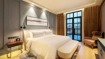 A modern hotel room with a large bed, a bedside table with a radio, a chair, and a view of the outdoors through a large window.
