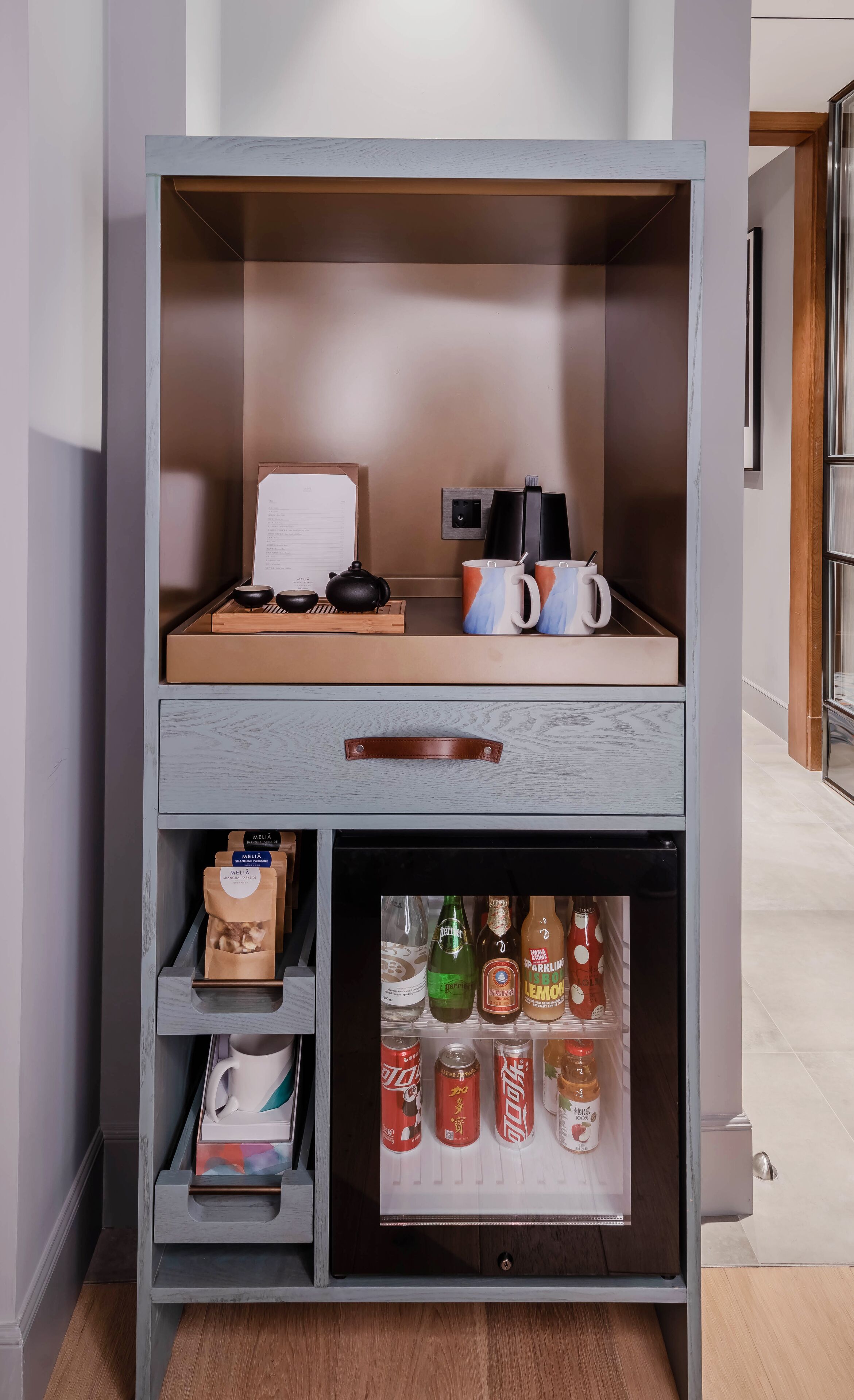 Un meuble de rangement compact doté d’une étagère supérieure pour une bouilloire, des tasses et un plateau, et d’une étagère inférieure pour des bouteilles et des canettes de boissons.