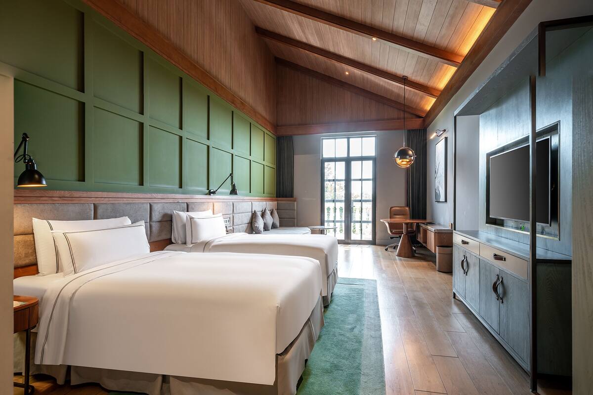 A hotel room with two large beds, a desk, a chair, a television, and a large window.