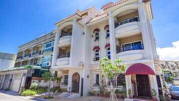 A multi-story building with balconies, a central entrance, and a small garden area with potted plants.