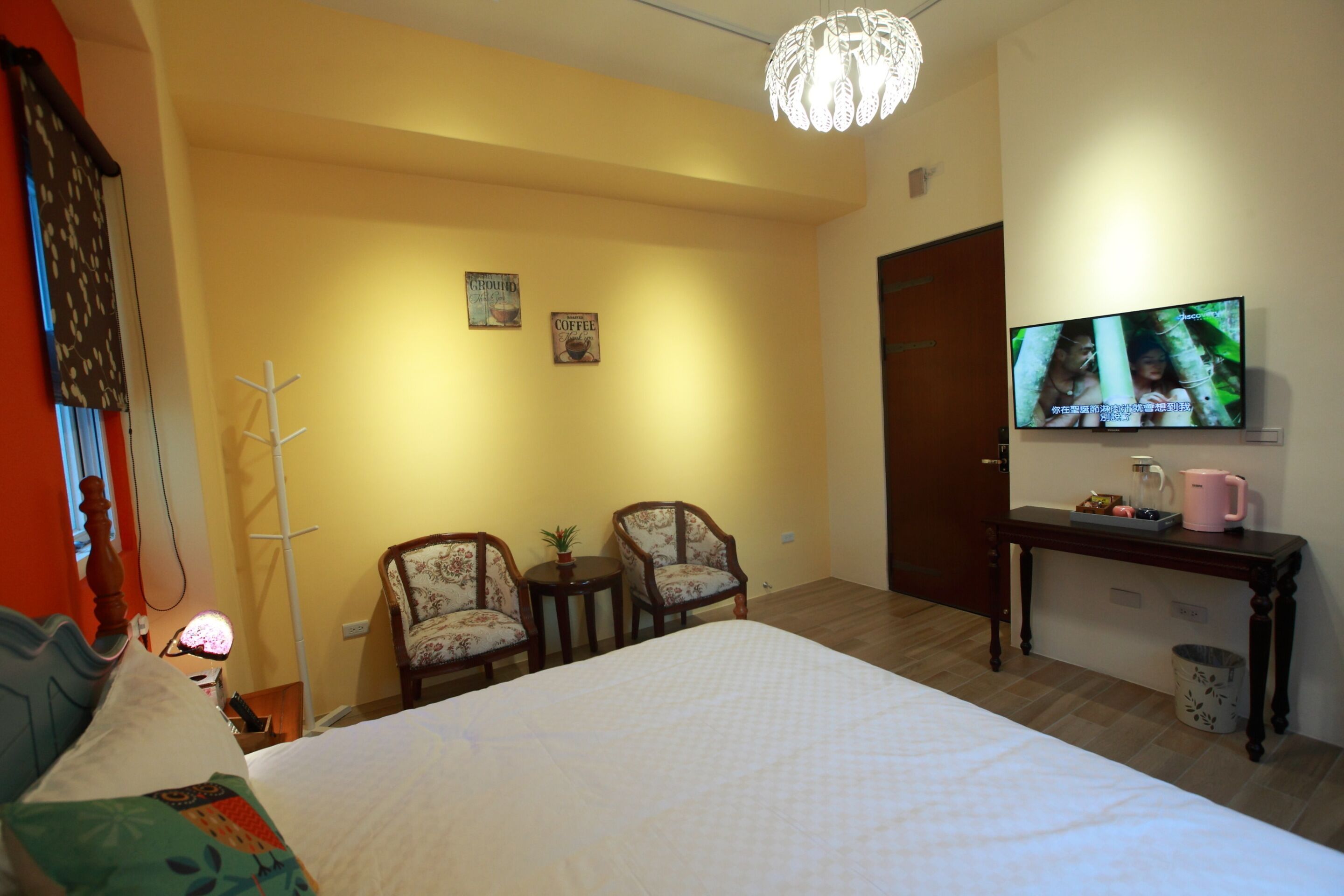 A hotel room with a bed, two chairs, a small table with a plant, a television on the wall, and a wooden desk with a pink mug.