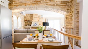 A dining area with a brick wall, a wooden table set for two, and a view of the kitchen in the background. - Anesis Family Villas (Faistos)