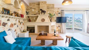 A living room with a stone fireplace, a wooden coffee table, and a large window offering a view of the city. - Anesis Family Villas (Faistos)