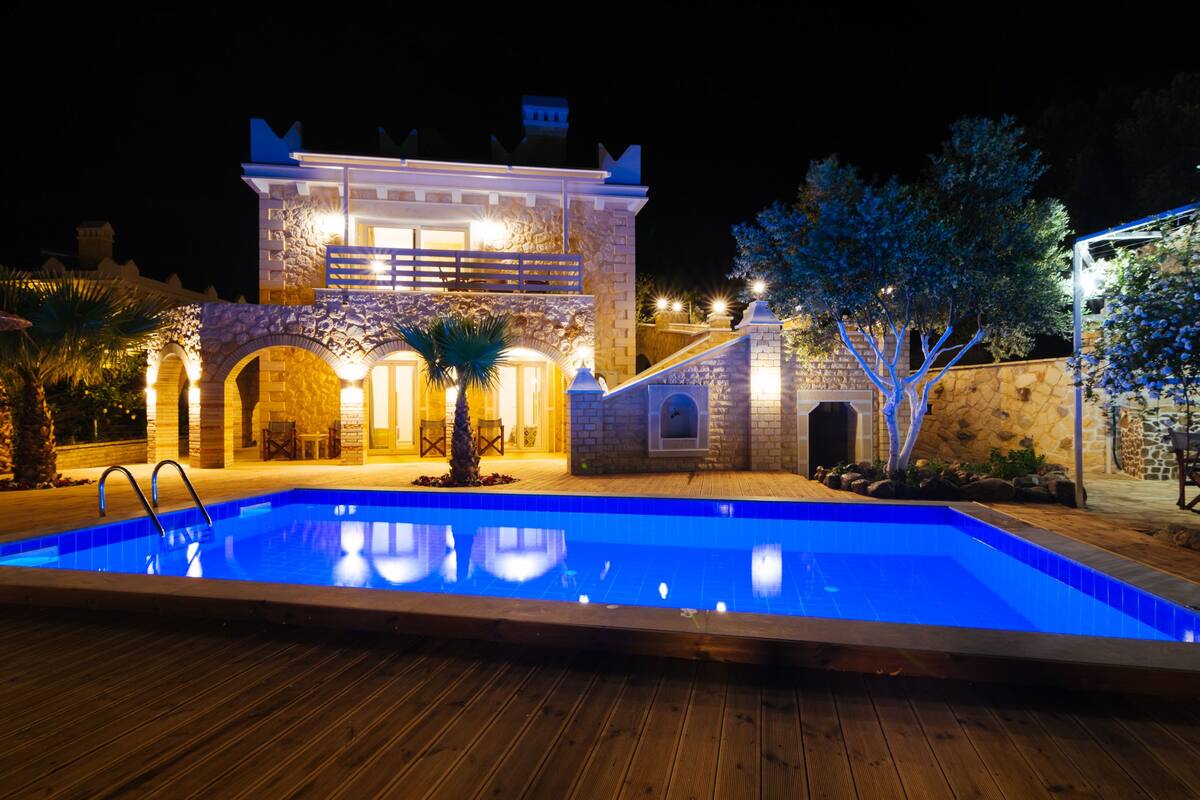 A swimming pool with a wooden deck, a stone building with arches and a balcony, and a tree with blue lights.