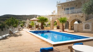 A pool area with wooden decking, lounge chairs, and umbrellas. - Anesis Family Villas (Faistos)