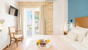 A hotel room with a bed, a TV mounted on the wall, a chair, a table with breakfast items, and a view of the outside. - Anesis Family Villas (Faistos)