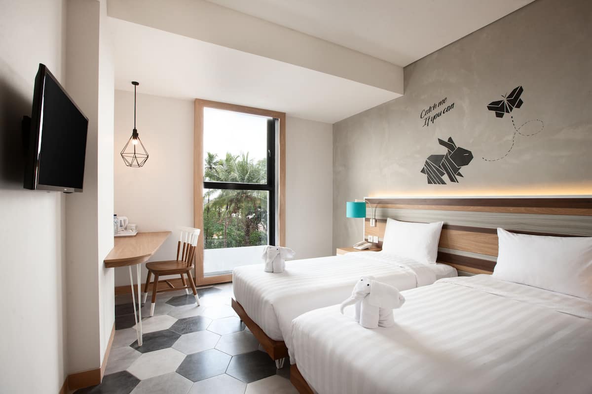 A hotel room with two beds, a desk, a TV, and a window with a view of trees.