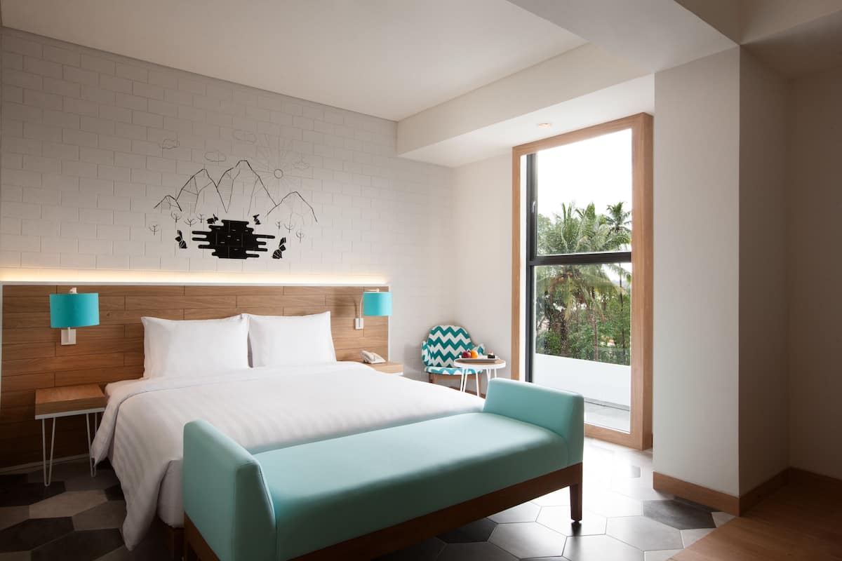 A modern hotel room with a large bed, a wooden headboard, a wall mural, a small table with a chair, and a large window offering a view of palm trees.