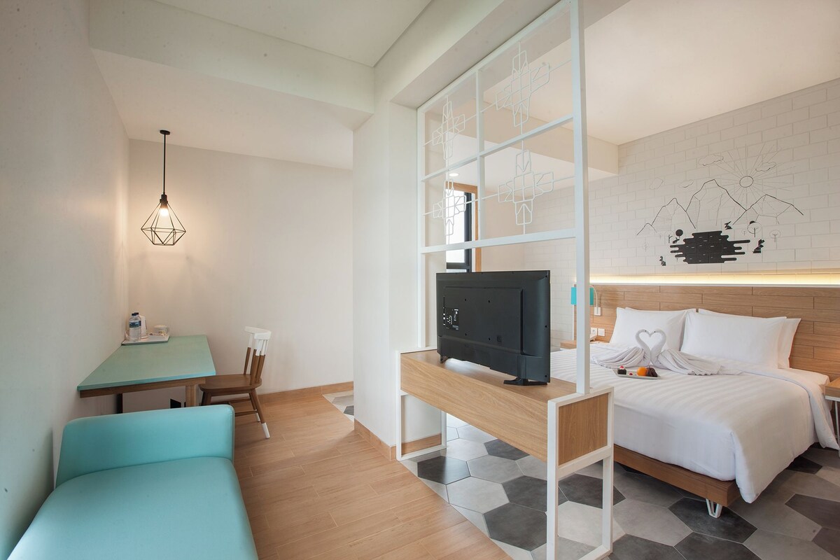 A modern hotel room with a bed, a desk with a television, a seating area, and a wall art piece.