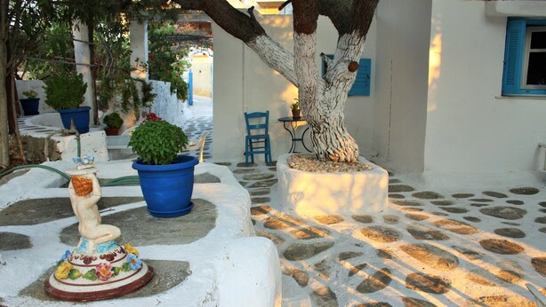 A courtyard with a tree, potted plants, a statue, and a stone floor. - Meltemi Studios and Apartments (Paros)