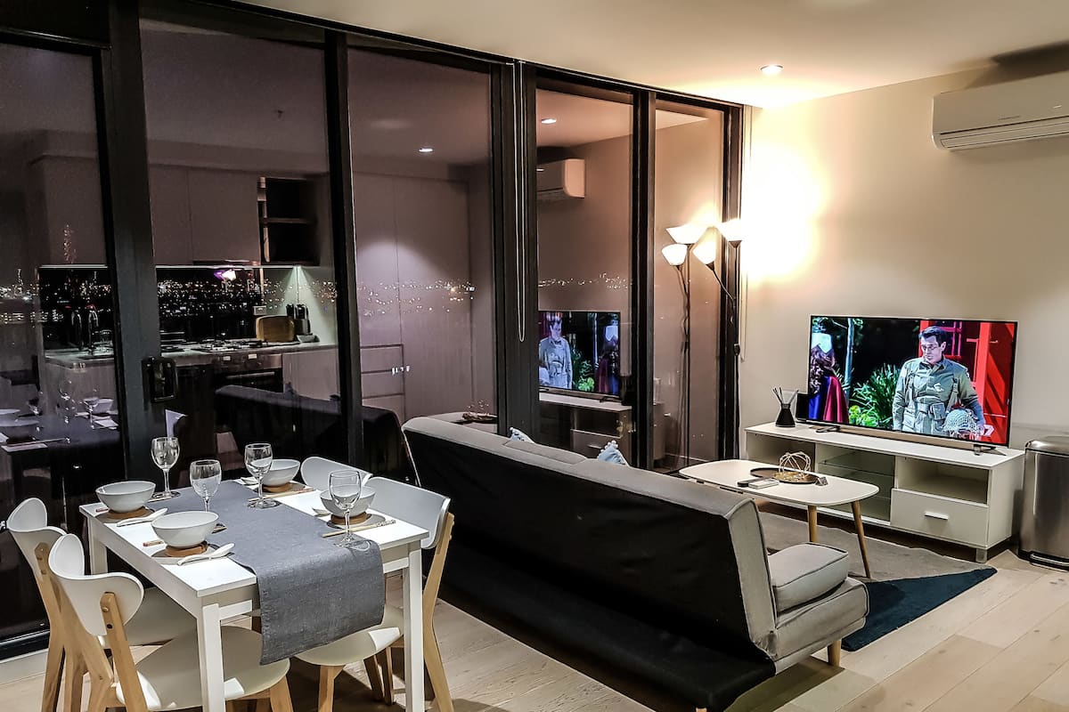 Un comedor moderno con una mesa para dos, un sofá y un televisor mostrando un programa.