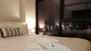 Een net opgemaakt bed met witte lakens en een opgevouwen handdoek, een gestreept kussen en een nachtkastje met een lamp.
