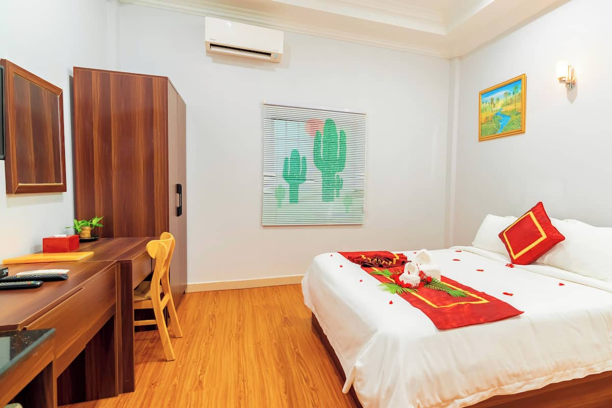 Une chambre d’hôtel avec un bureau en bois, un lit avec une literie blanche et des touches de couleur rouge, un tableau au mur et un appareil de climatisation.