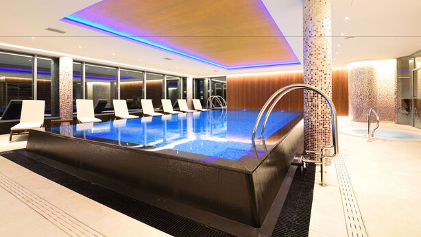 Una piscina interior moderna de forma larga y estrecha, rodeada de sillones de descanso y un muro de azulejos.