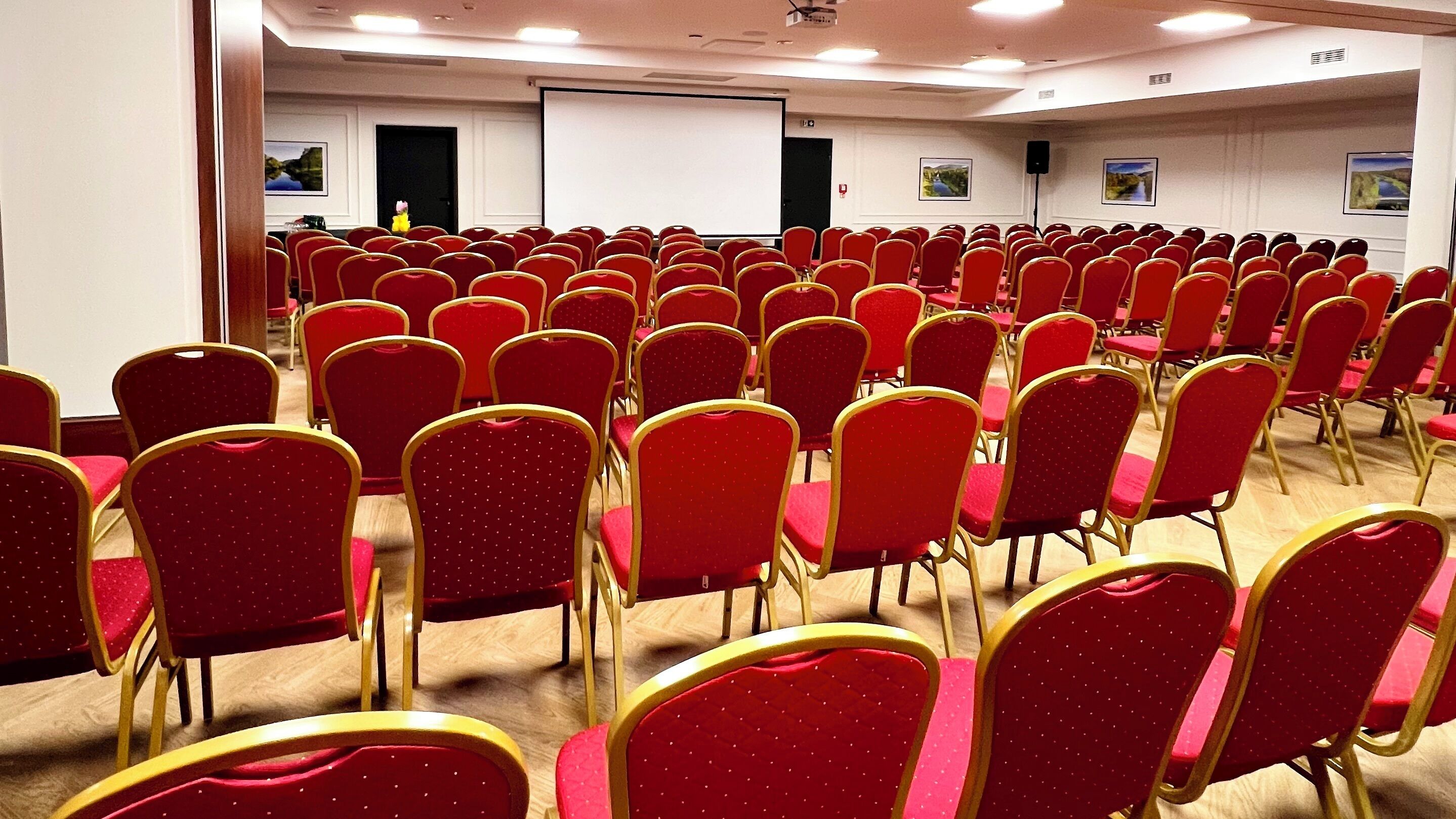 Un salón de conferencias con filas de sillas rojas y una pantalla para proyector.