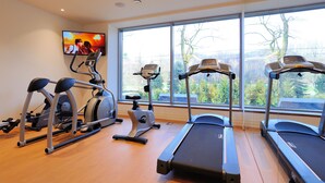 Un gimnasio con cintas de correr, elípticas y una bicicleta estática, amplias ventanas con vistas a árboles, y un televisor en la pared.