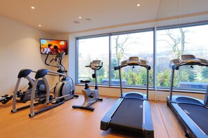 Un gimnasio con cintas de correr, elípticas y una bicicleta estática, amplias ventanas con vistas a árboles, y un televisor en la pared.