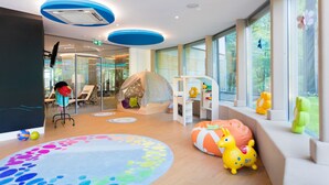 Un área de juegos infantil moderna con un ventanal grande, una carpa de juegos y coloridos pufs de relleno.